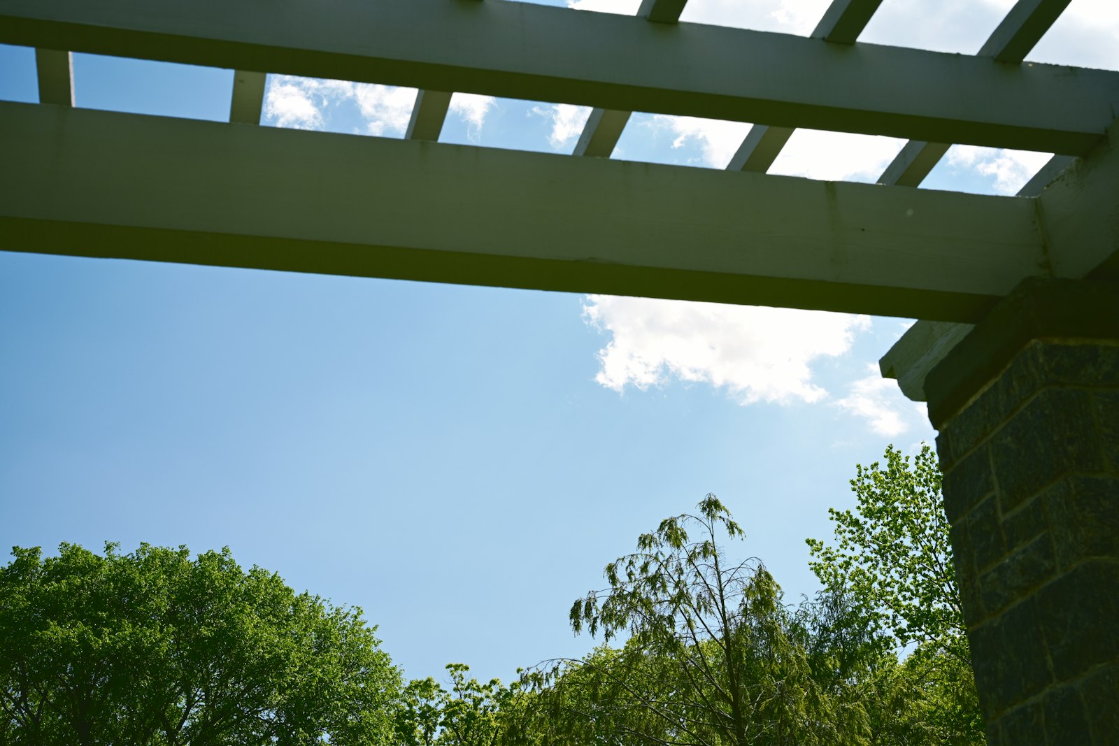 Looking up at the open wood beams of a pergola against a blue, partly cloudy sky.